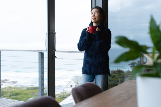 Asian woman holding mug of coffee and leaning on window