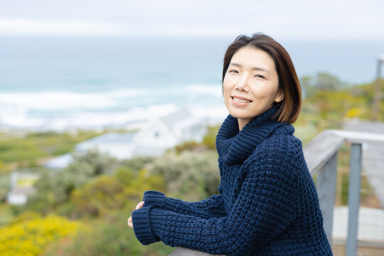 Portrait of happy asian woman wearing jumper and leaning on balcony alone