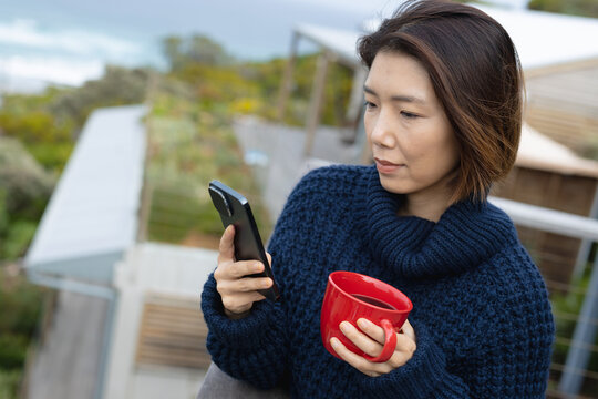 Asian woman holding mug of coffee and using smartphone