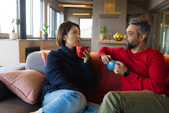 Happy diverse couple sitting on sofa in living room, holding mugs