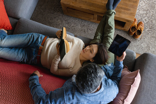 Happy diverse couple sitting on sofa in living room, using tablet and reading book
