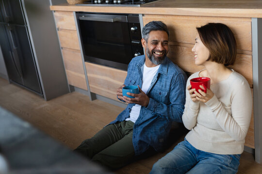 Happy diverse couple sitting on floor in kitchen, holding mugs