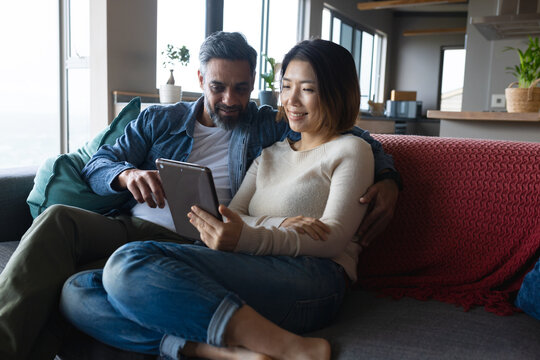 Happy diverse couple sitting on sofa in living room, using tablet