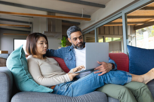 Happy diverse couple sitting on sofa in living room, using laptop