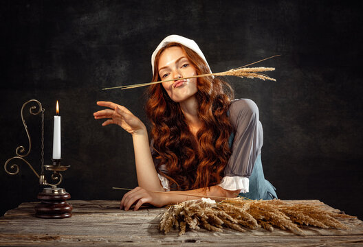Creative portrait of young beautiful redhead girl with long curly hair in image of medieval person in renaissance style dress isolated on dark background.