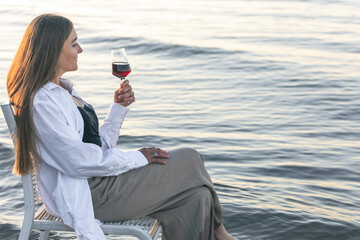 A beautiful woman with a glass of wine on the seashore sits on a chair.