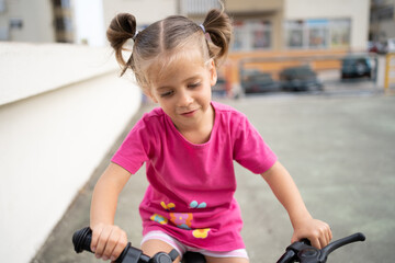 Cute little smiling girl riding bike bicycle in city on parking sunny summer day. Active family leisure with kids.