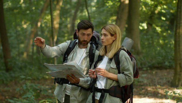 Couple of active middle aged tourists using paper map to find right route, discussing way during hiking trip in forest