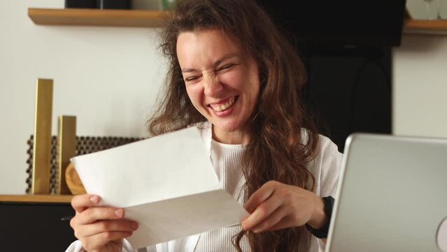 Happy young caucasian woman reading papper letter about college admission, scholarship approved, invitation for internship abroad,sit with laptop at home.Got hired job of dream,celebration of victory