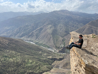 Tourist sits on edge of high mountain cliff and admires mountains of North Caucasus, Goor village area in Dagestan