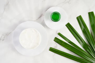 A mock-up of a round cosmetics container with cream on marble background and green palm leaf, top view