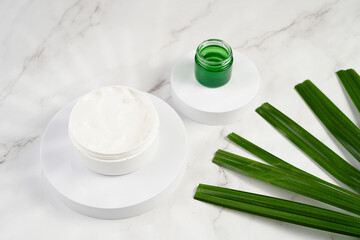A mock-up of a round cosmetics container with cream on marble background and green palm leaf, top view