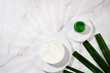 A mock-up of a round cosmetics container with cream on marble background and green palm leaf, top view