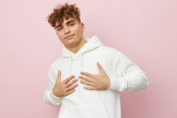 a handsome, pleasant young man stands on a pink background in a white hoodie and looks at the camera smiling pleasantly with his hands on his chest. Horizontal Studio Photography