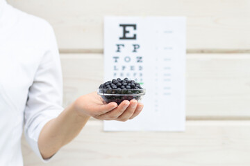A bowl of blueberries in a doctor's female hand, Snellen's vision chart in the background. Natural vitamins for the eyes.
