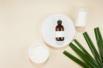 A mock-up of a brown bottle with pipette, white cosmetics container and container with cream on white round podium on beige colored background, green palm leaf,top view, space for text