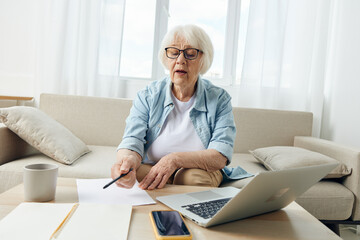 an elderly woman is passionate about the workflow, sitting at a coffee table on a cozy sofa at home and holding a training video conference, pointing with a pen to a sheet of paper with notes