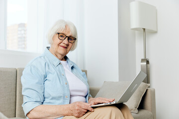 Fototapeta na wymiar a happy, carefree elderly woman with gray hair is sitting on the couch working from home in a cozy apartment, holding a laptop on her lap and smiling pleasantly while looking at the camera