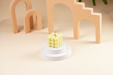 Pale green bubble candle burning on two white round podiums in peach colored arched doors for cosmetics photography on beige colored background