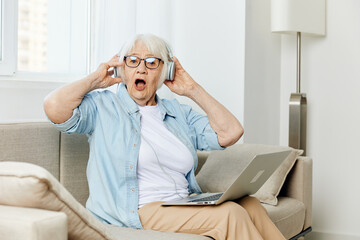 Fototapeta premium a pleasant joyful elderly lady uses modern technologies holding a laptop on her lap and holding headphones with her hands
