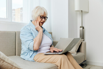 an elderly lady in stylish clothes is sitting on a cozy beige sofa looking at a laptop monitor that she holds on her lap and emotionally spreads her hands