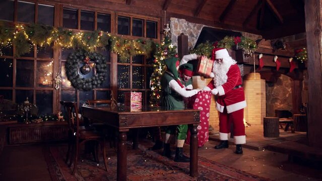 Santa's residence at Christmastime. Elves and Santa Claus pack red sack with gift boxes for kids in a beautiful Christmas decorated residence.