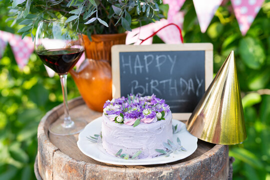 A very beautiful bento cake with purple, flowers on a background of pink flags. Birthday. In the background, a bouquet of flowers, a jug of flowers, a cap for the head, a board with an inscription
