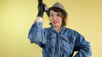 Forty-year-old woman in a denim jacket and hat shoots with a pistol on an isolated yellow background. Portrait of a bandit woman - Powered by Adobe