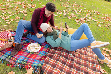 Fototapeta premium Happy biracial couple having picnic on a rug and using smartphone in autumn garden, smiling