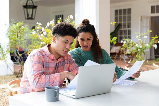 Biracial couple sitting on garden terrace outside house using laptop and paying bills