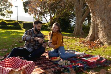 Fototapeta premium Happy caucasian couple having picnic sitting on rug in sunny autumn garden pouring wine