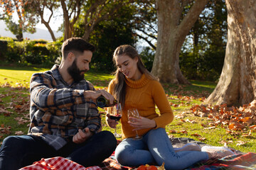 Fototapeta premium Happy caucasian couple sitting on rug in sunny autumn garden drinking wine