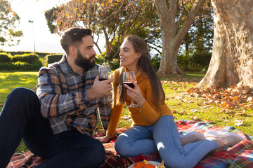 Fototapeta premium Happy caucasian couple sitting on rug in sunny autumn garden drinking wine