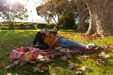 Fototapeta premium Happy caucasian couple lying on rug having picnic in sunny autumn garden and drinking wine