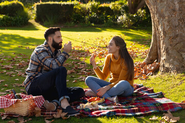 Fototapeta premium Happy caucasian couple sitting on rug having picnic and talking in sunny autumn garden