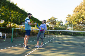 Fototapeta premium Happy caucasian couple playing tennis making a toast with water bottles on outdoor tennis court