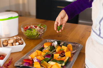 Midsection of caucasian woman standing in kitchen sprinkling fresh herbs on chopped vegetables