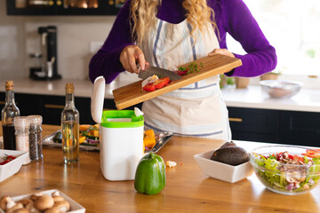 Midsection of caucasian woman standing in kitchen in apron composting vegetable scraps