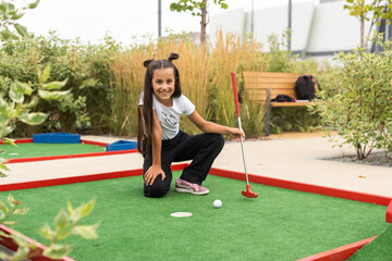 Miniature golf outdoor. Little caucasian girl golfing in the mini golf course. - obrazy, fototapety, plakaty