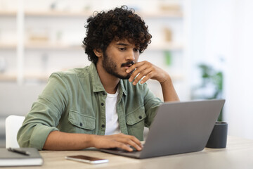 Pensive dark-skinned guy sitting at workdesk in front of laptop