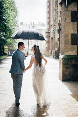 Stylish bride and groom walking under umbrella and kissing on background of old church in rain. Provence wedding. Beautiful wedding couple embracing together in rainy street. Back view