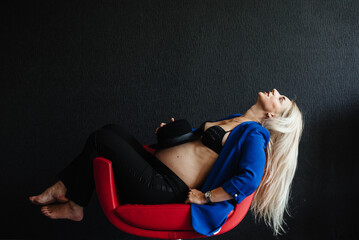 Pregnant woman in a blue jacket and pants sitting in a stylish red chair on a background of black...