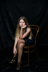 sad beautiful young blonde girl with long hair in shining blue dress sitting on the chair on  black background