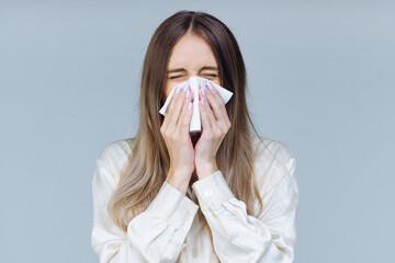Studio portrait of cute unhealthy Caucasian female with paper napkin sneezing, experiences allergy symptoms, caught a cold