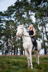 young beautiful blond smiling woman with long hair in black dress riding a white horse with blue eyes in autumn field	