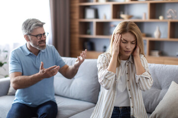 Sad upset angry adult european husband yells at unhappy wife, covers ears in living room interior - obrazy, fototapety, plakaty