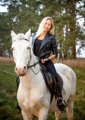 young beautiful blond smiling woman with long hair  riding a white horse with blue eyes in autumn field	