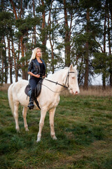 young beautiful blond smiling woman with long hair in black dress riding a white horse with blue eyes in autumn field	