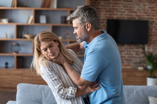 Aggressive angry european husband strangles behind proud frightened afraid wife in living room interior