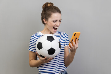 Portrait of excited positive happy woman wearing striped T-shirt holding soccer ball and smart...